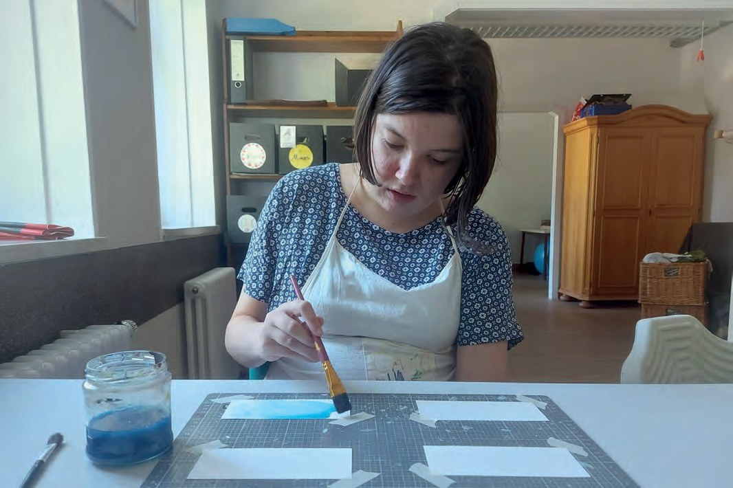Eine junge Frau mit Schürze malt mit einem Pinsel und blauer Farbe auf Papier, das auf einer Schneidematte festgeklebt ist.