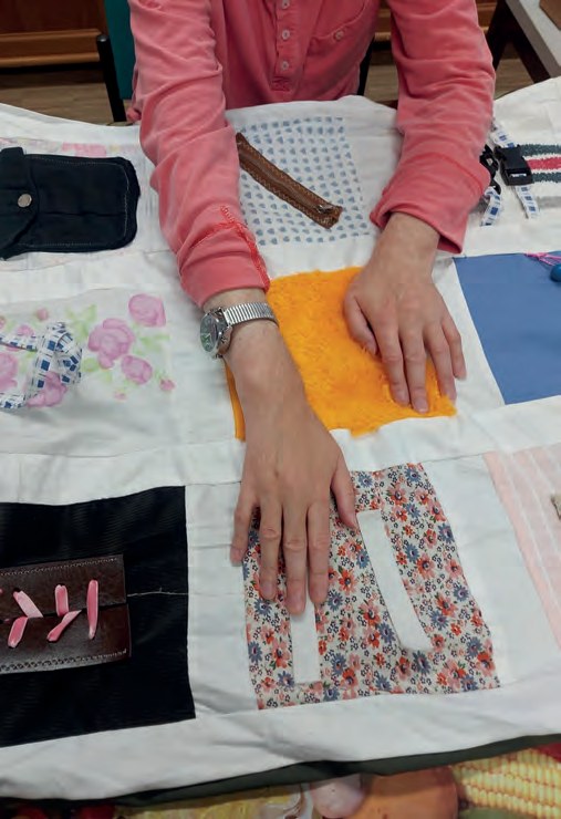 Hände legen bunte Stoffstücke auf einer Patchwork-Decke zurecht, darunter Jeansstoff, geblümte Stoffe und ein gelbes Filzstück.