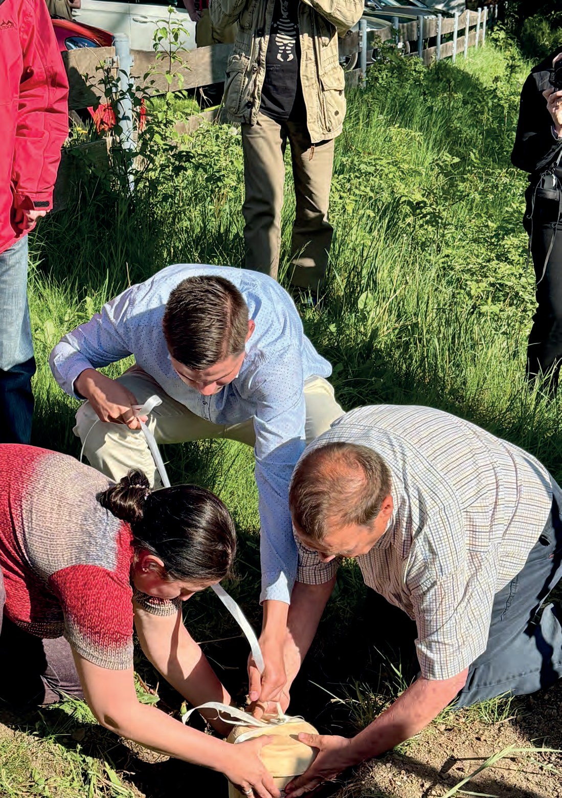 Drei Menschen knien im Gras und setzen gemeinsam den Grundstein in die Erde, dahinter stehen Gäste.