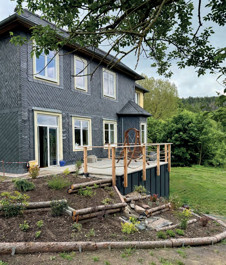 Ein Haus mit dunkler Schieferfassade und einer neuen Holzterrasse, davor ein frisch angelegtes Beet mit Holzstämmen als Einfassung.