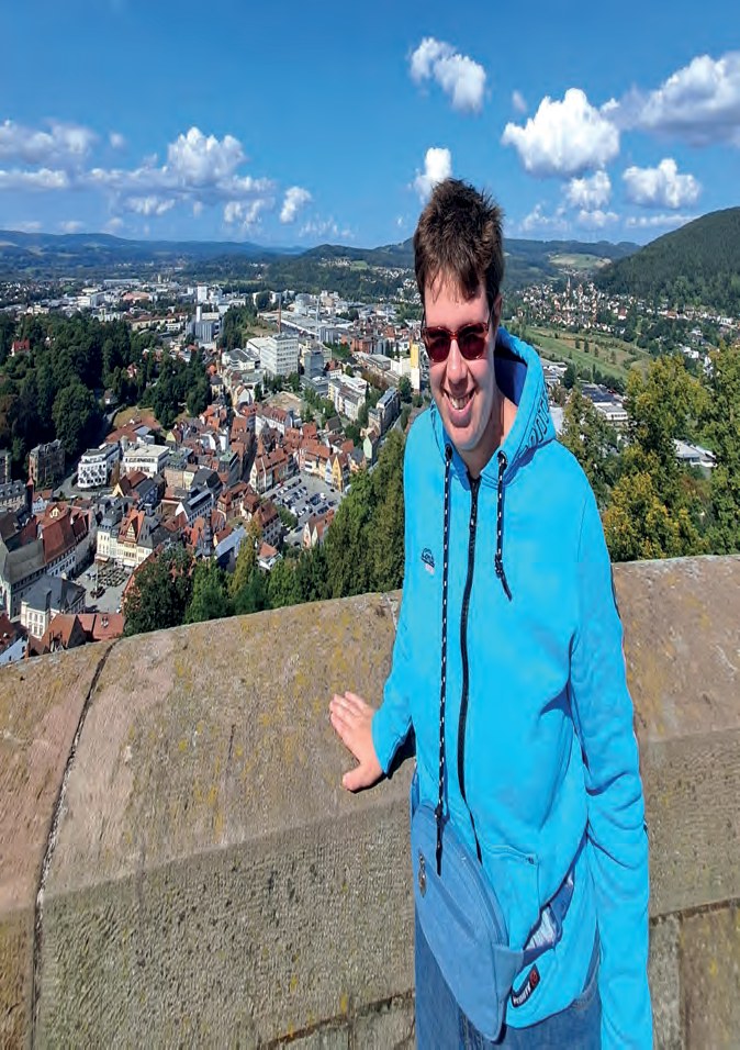 Jasmin Hügerich in einer hellblauen Jacke und mit Sonnenbrille steht auf einer Aussichtsmauer, hinter ihr der Blick über eine Stadt im Tal und bewaldete Hügel.