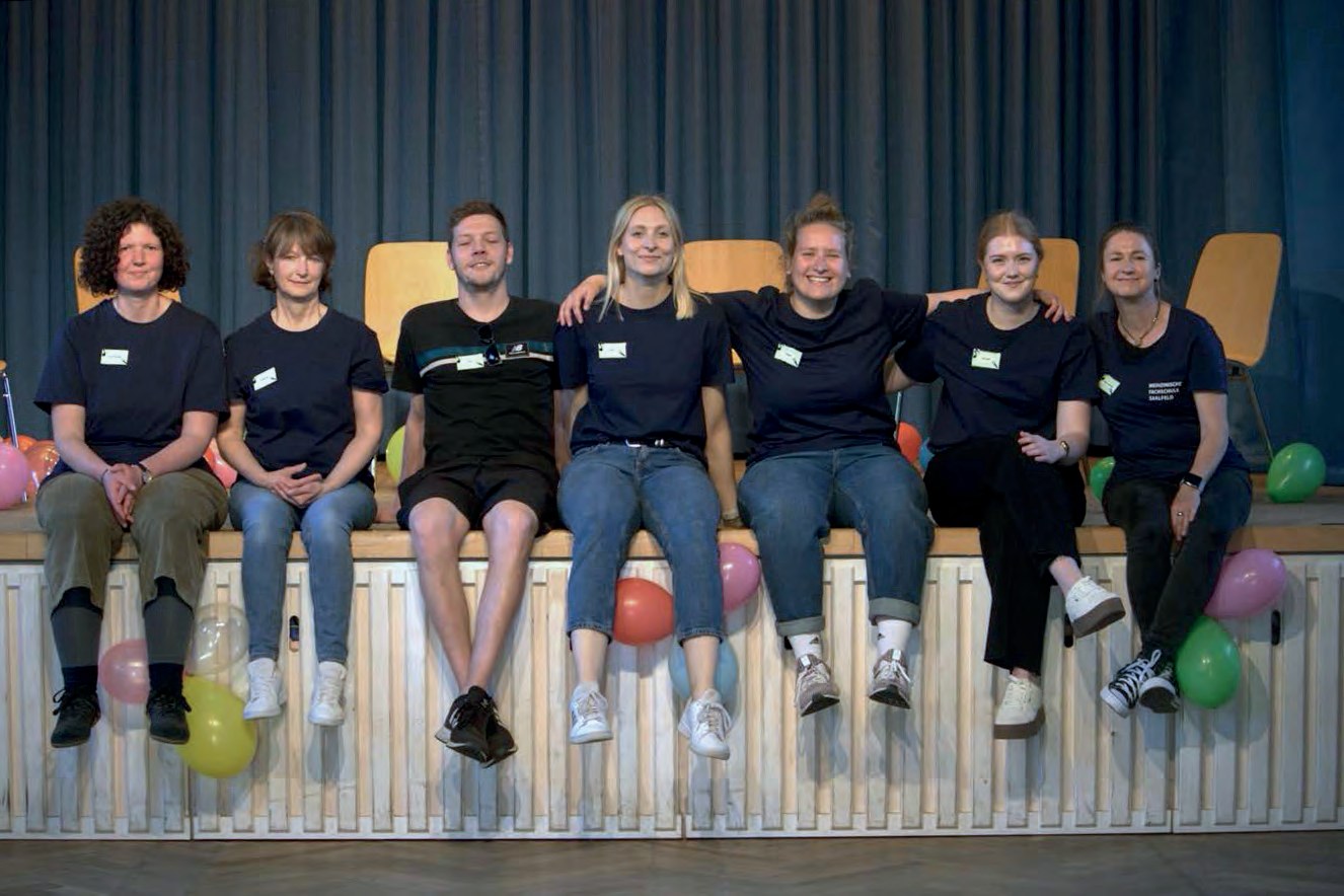 Sieben junge Frauen und Männer in dunkelblauen T-Shirts sitzen nebeneinander auf dem Bühnenrand, um sie herum Luftballons.