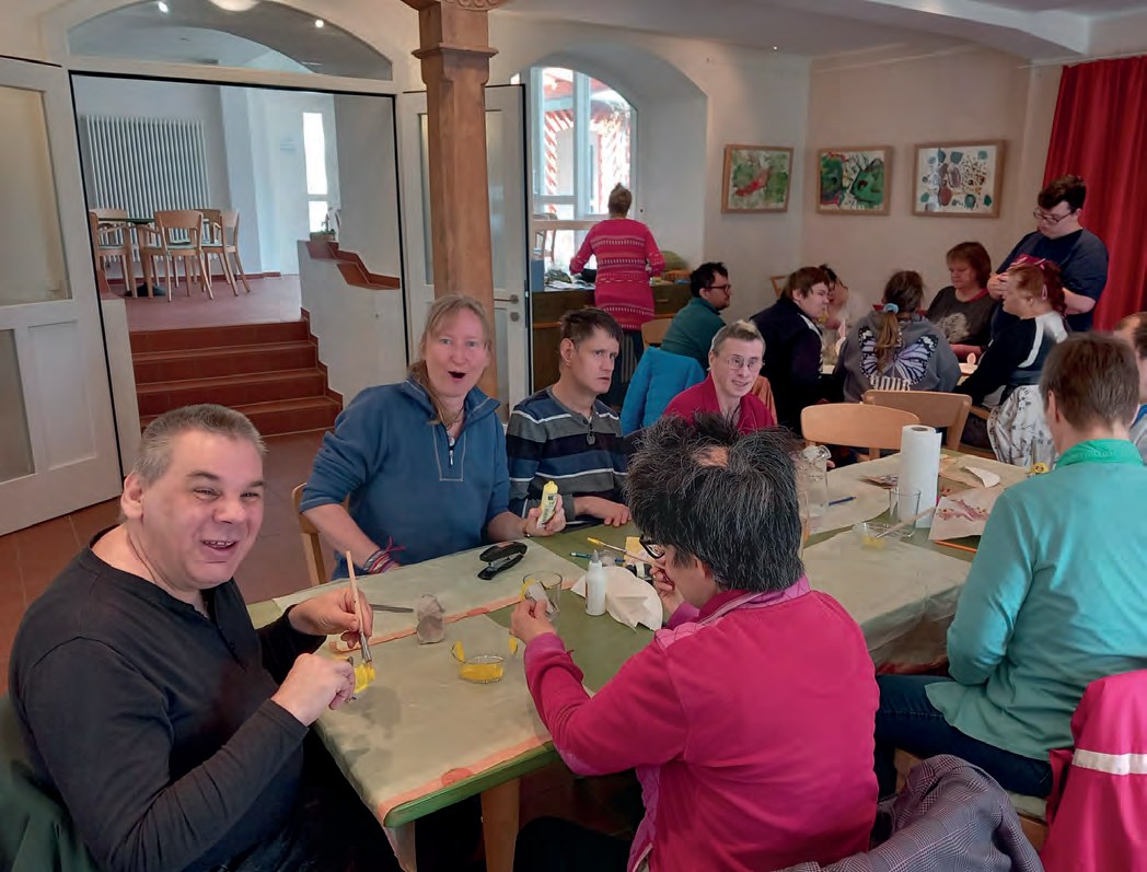 Weitere Tische in der Cafeteria: Bewohner basteln mit Pinseln und gelben Papierküken, ein Mann im Vordergrund lacht in die Kamera.