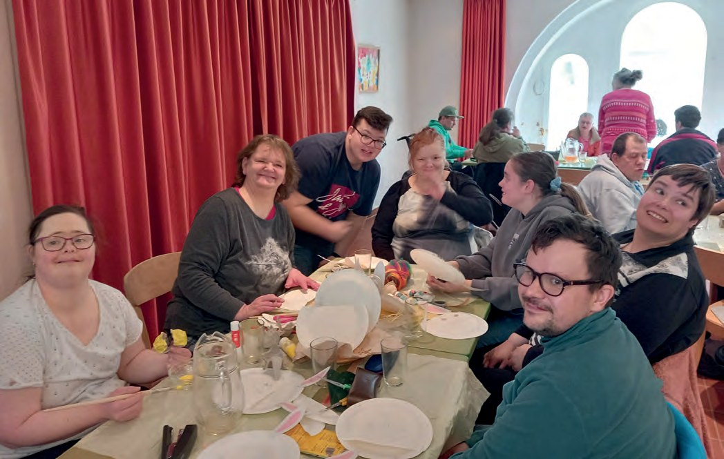 Bewohnerinnen und Bewohner sitzen fröhlich an einem langen Tisch voller Pappteller, Kleber und Bastelsachen. Einige lachen in die Kamera.