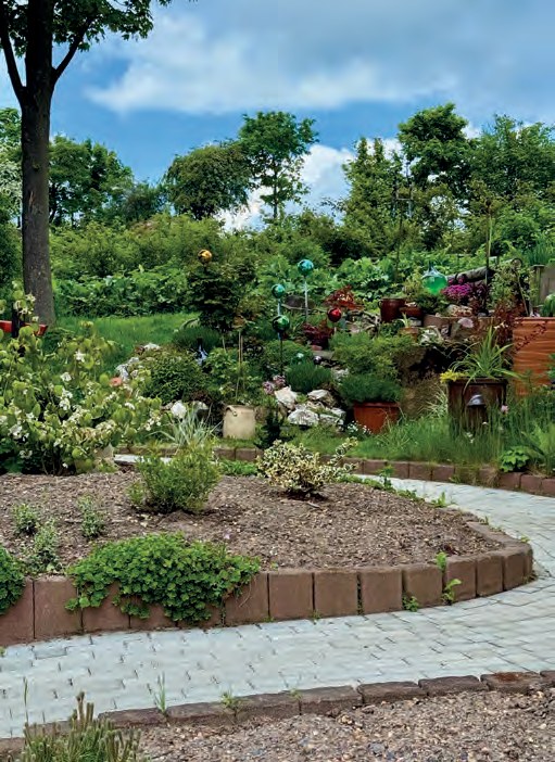 Ein neu angelegter Garten auf einer Anhöhe: geschwungene Beete mit Steinkanten, Blumen und Sträuchern, gepflasterte Wege, dahinter Bäume und blauer Himmel.