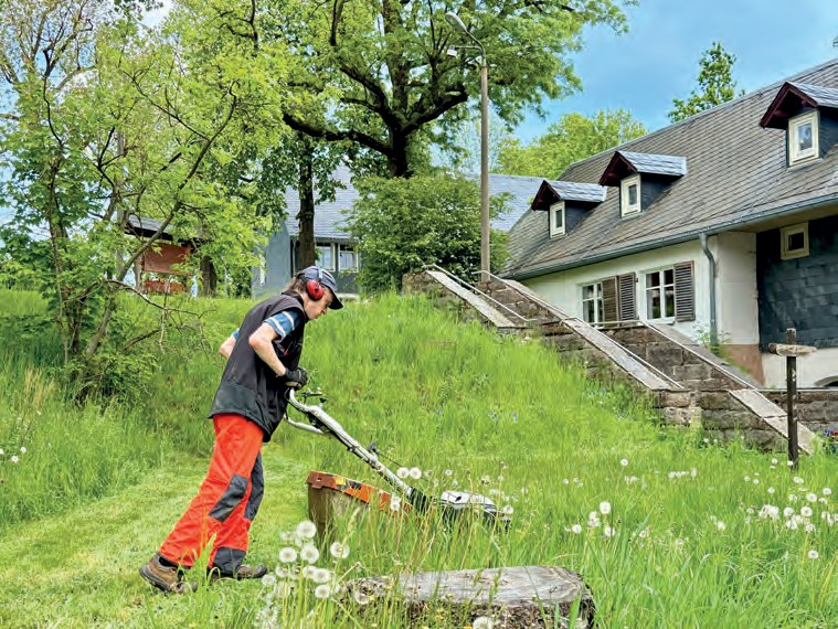 Ein Mann mit Gehörschutz und orangefarbener Arbeitshose mäht eine Wiese voller Pusteblumen, dahinter ein Haus mit Schieferdach und eine Treppe.