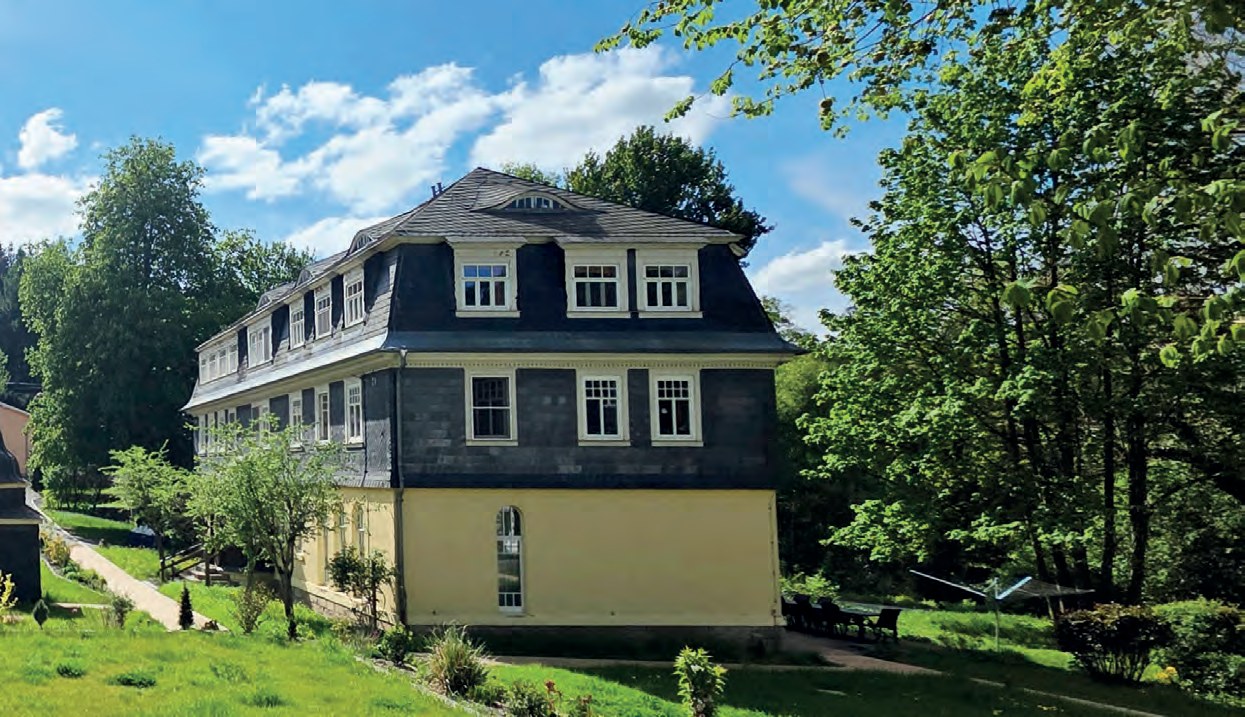 Ein großes Haus mit gelbem Erdgeschoss und dunkler Schieferfassade im Grünen, davor eine frisch angesäte Rasenböschung, junge Sträucher und hohe Laubbäume.