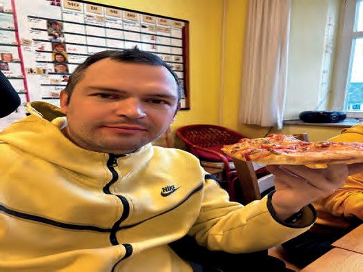 Ein Bewohner in gelber Trainingsjacke hält ein Stück Pizza in die Kamera.