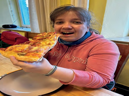 Eine Bewohnerin im rosa Pullover hält lachend ein großes Stück Pizza in beiden Händen.